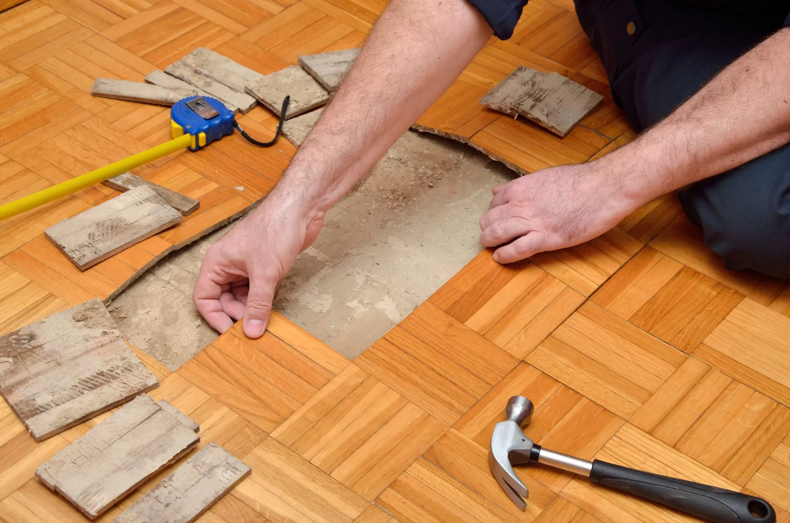 A hand places a replacement wooden tile on a floor, surrounded by measuring tape and tools, during a Wood Floor Repair